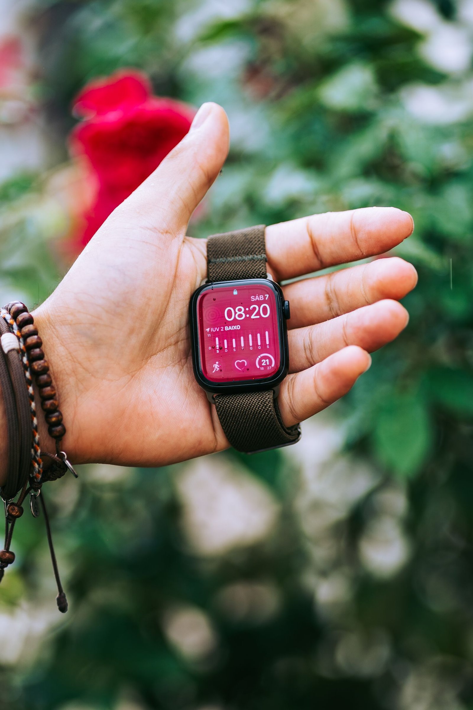 Close-up of a hand holding a smartwatch with a red display and green strap, set against a blurred outdoor background.