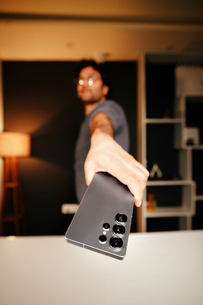A man holds a smartphone towards the camera in a stylish indoor setting, showcasing device features.