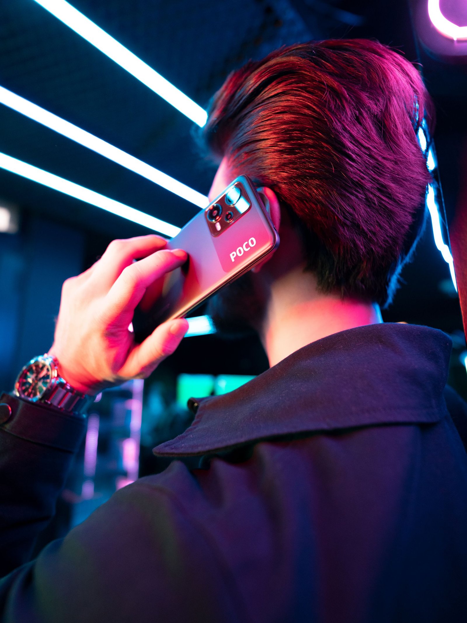 A man with short hair holds a smartphone in a neon-lit setting, seen from behind.