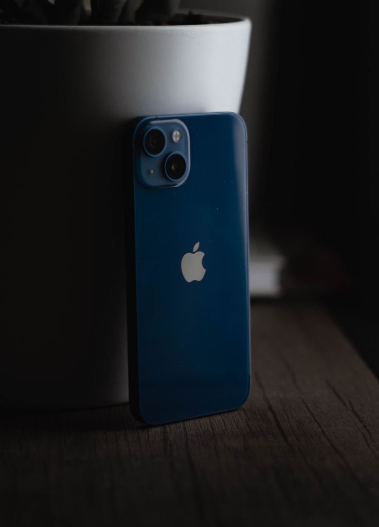 Blue smartphone leaning against a plant pot on a wooden surface, partially in shadow.